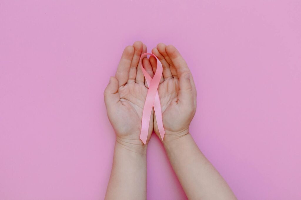 Child's hands holding pink ribbon for breast cancer awareness on pink background.